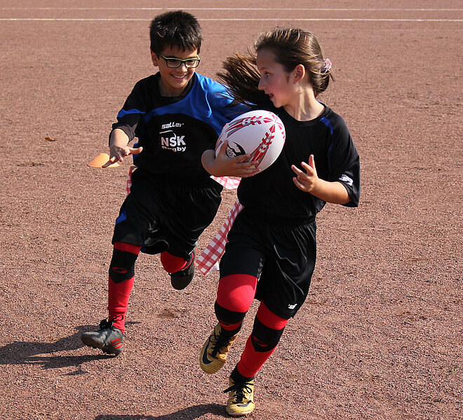 Zwei Kinder spielen mit einem Rugbyball auf einem Ascheplatz.