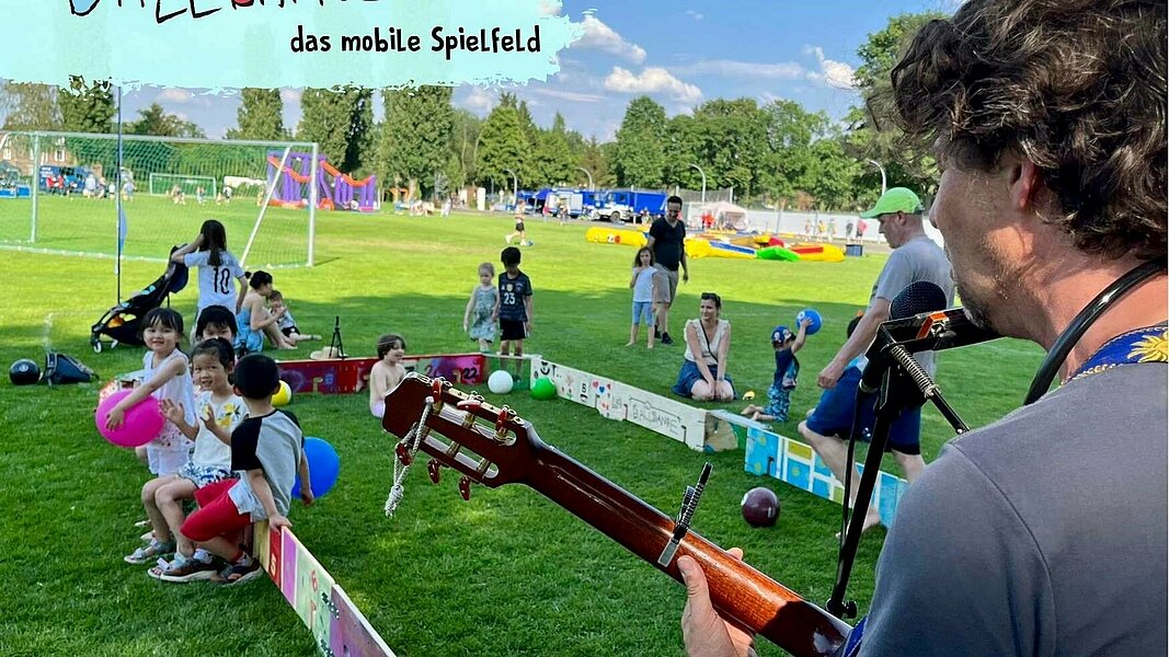 Mann spielt im Vordergrund Gitarre, Kinder spielen im Hintergrund mit der Ballbande und Bällen.