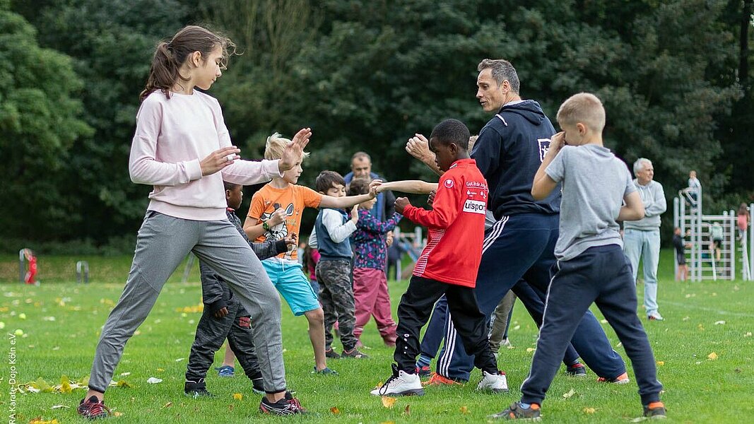 Eine Gruppe von Kindern trainiert Karate unter Anleitung auf einer Wiese.