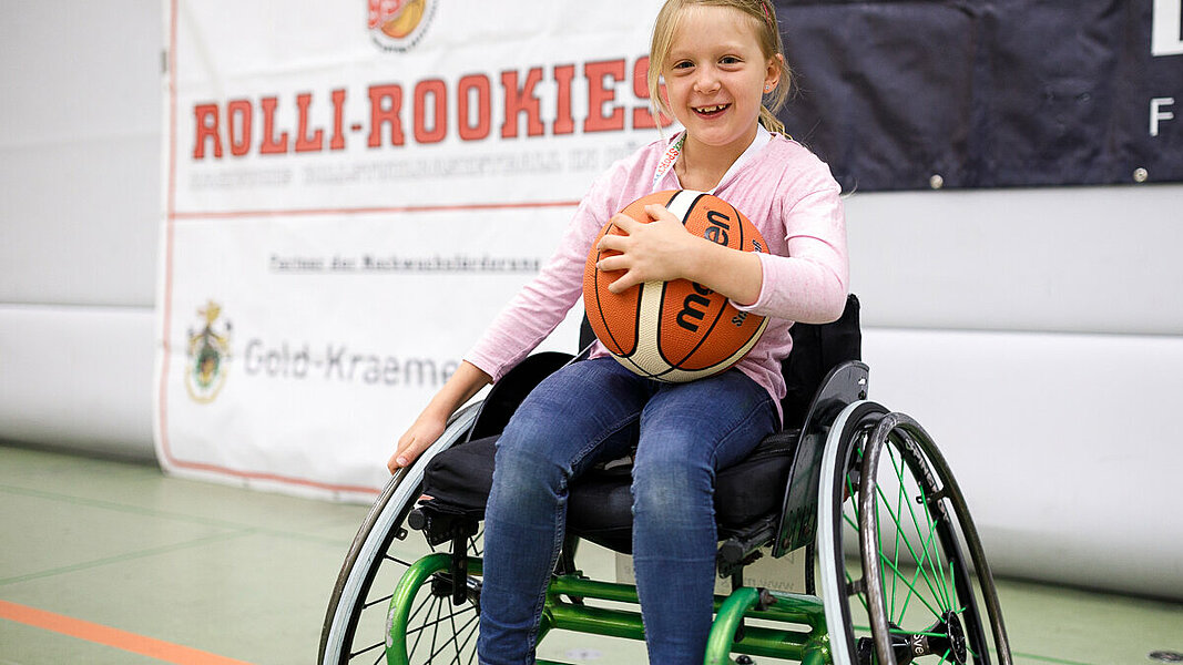 Mädchen in einem Sportrollstuhl mit einem Basketball in den Händen.