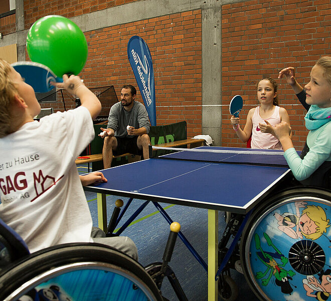 Kinder in Rollstühklen spielen Tischtennis in einer Sporthalle.