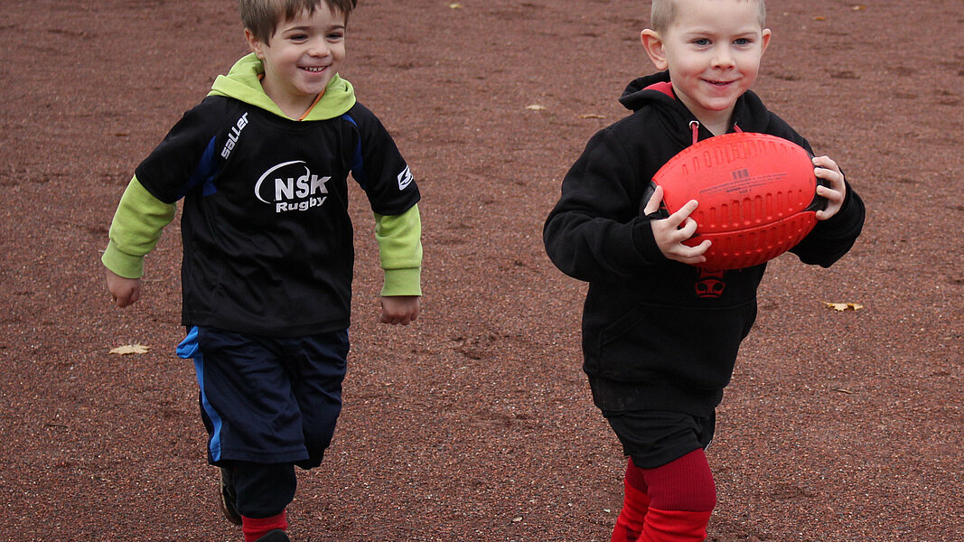 Zwei Kinder spielen mit einem Rugbyball auf einem Ascheplatz.