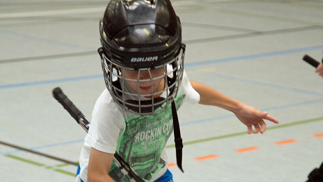 Junge mit Helm, Hockeyschläger und Inlineskate in einer Sporthalle.