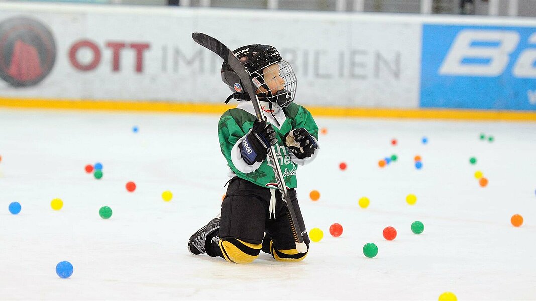 Ein Junge kniet in Esihockeymontur auf dem Eis und ist von bunten Bällen umgeben.
