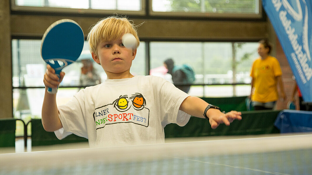 Ein Junge steht an einer Tischtennisplatte und will einen Tischtennisball schlagen.